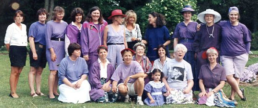 A group of 18 women and one young girl sit on a lawn dressed in purple, the LCIH staff.